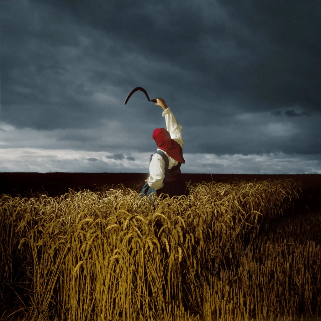 Woman wearing a red head covering carrying a scythe in a field