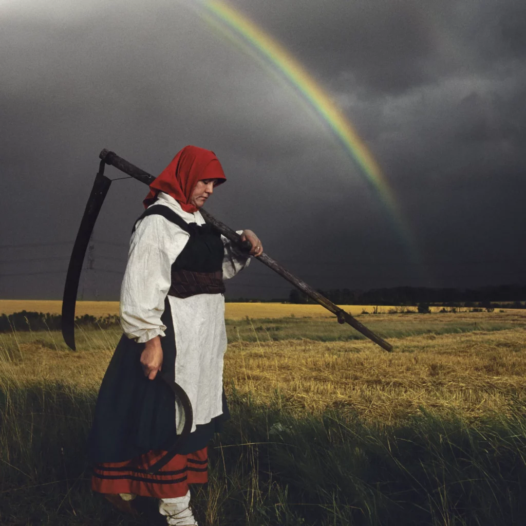 Woman wearing a red head covering carrying a scythe with a rainbow in the background